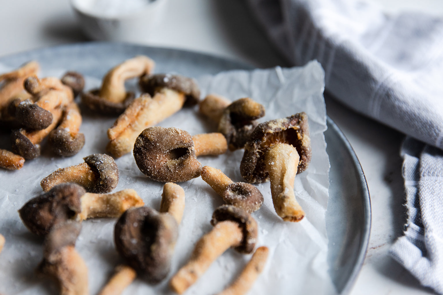 Fried bite-size mushrooms on a parchment-lined metal plate with a small bowl of salt and a cloth napkin in the background
