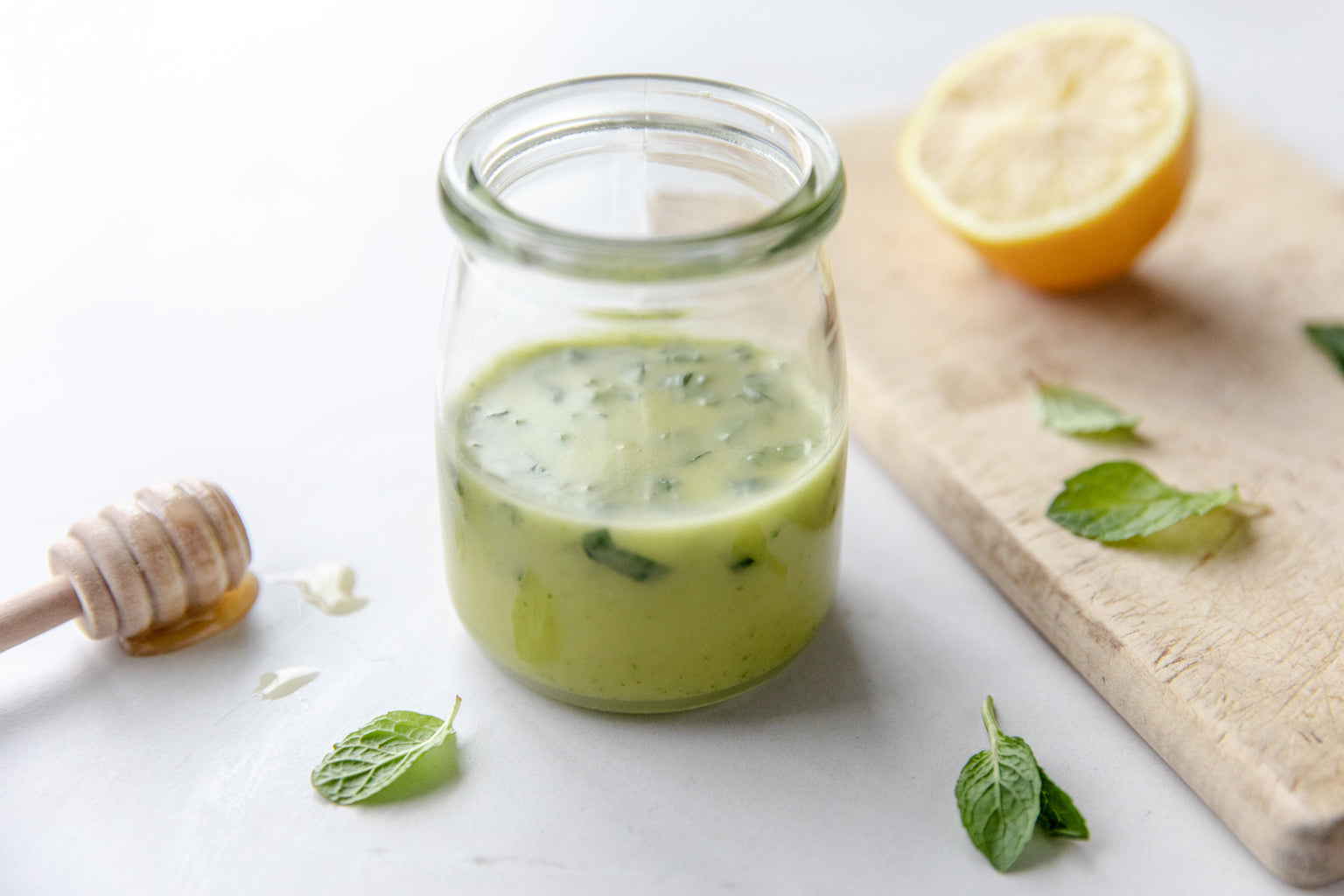 Jar of mint, honey, and lemon salad dressing on a white countertop with a halved lemon, scattered mint leaves, and a honey dipper