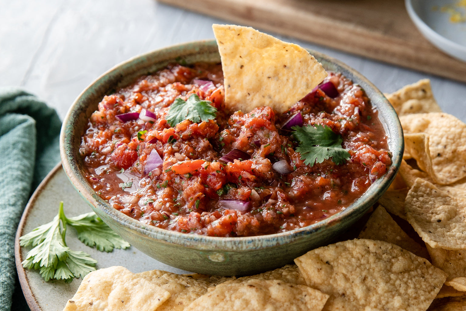Bowl of roasted red pepper and tomato salsa garnished with cilantro and red onion, tortilla chip dipped in salsa with extra chips on a plate