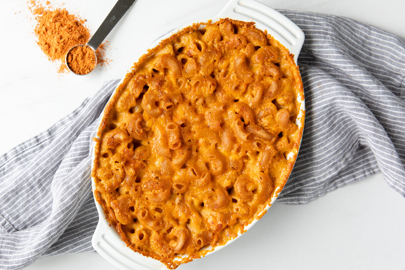 Overhead view of baked buffalo mac and cheese in an oval white baking dish on a gray striped cloth with a measuring spoon of buffalo seasoning