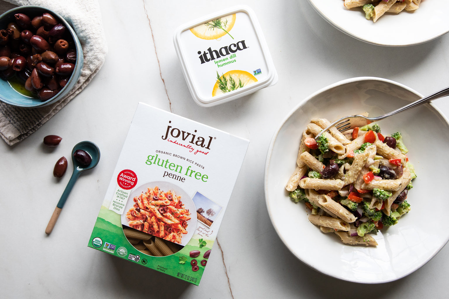 Bowl of hummus-dressed gluten-free penne pasta salad with olives beside Jovial gluten free penne box and Ithaca hummus