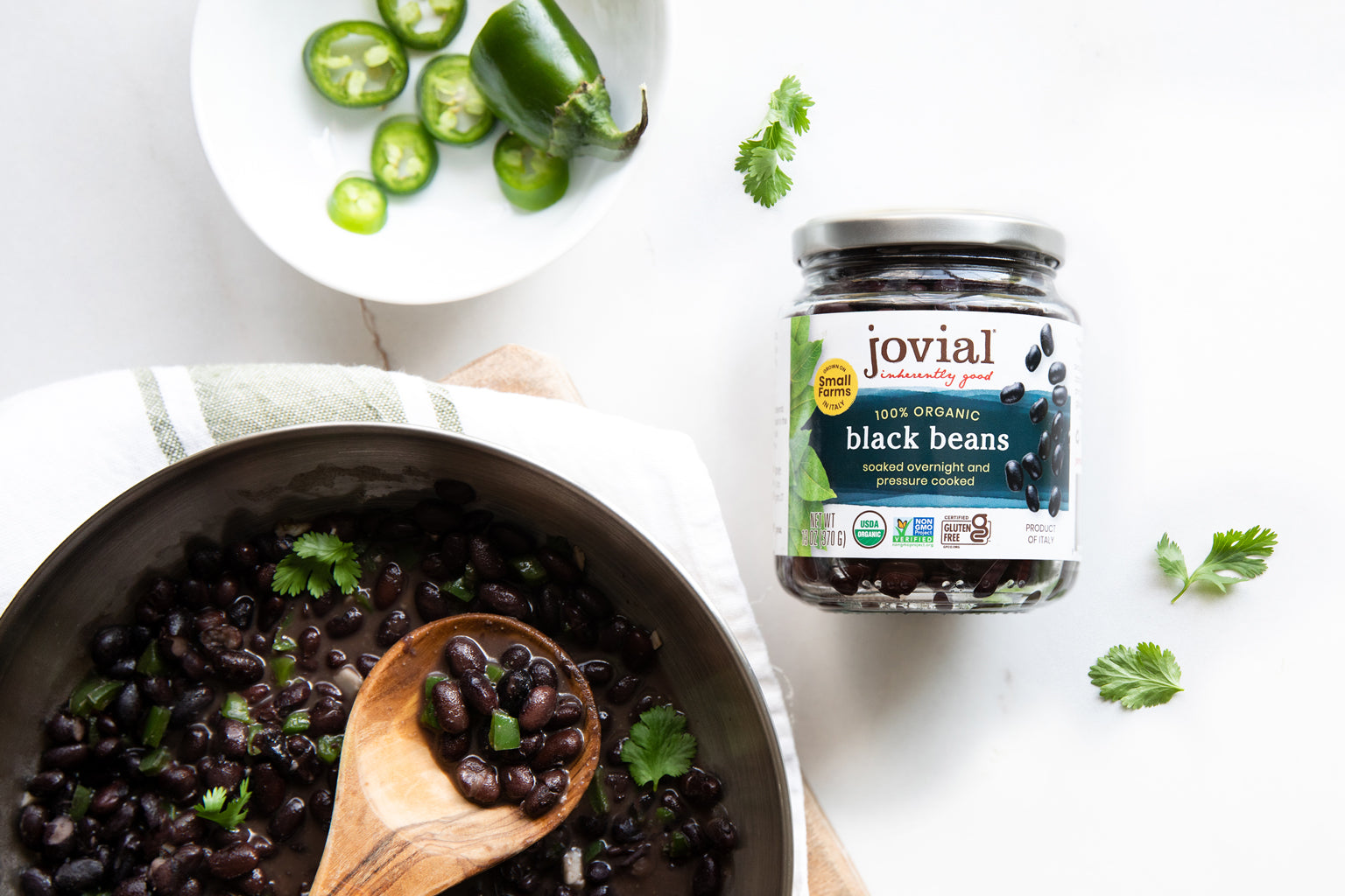 Overhead view of a Jovial black beans jar and a bowl of cooked beans showing the jar label and jalapeno and cilantro garnish on white
