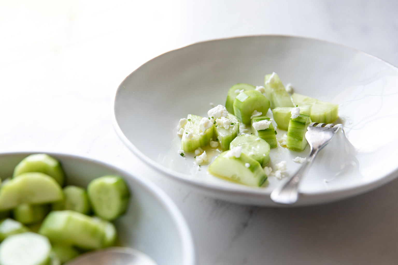 Cucumber and goat cheese salad served in a white shallow bowl with lemon and Jovial extra virgin olive oil, fork resting at the rim on a light marble surface