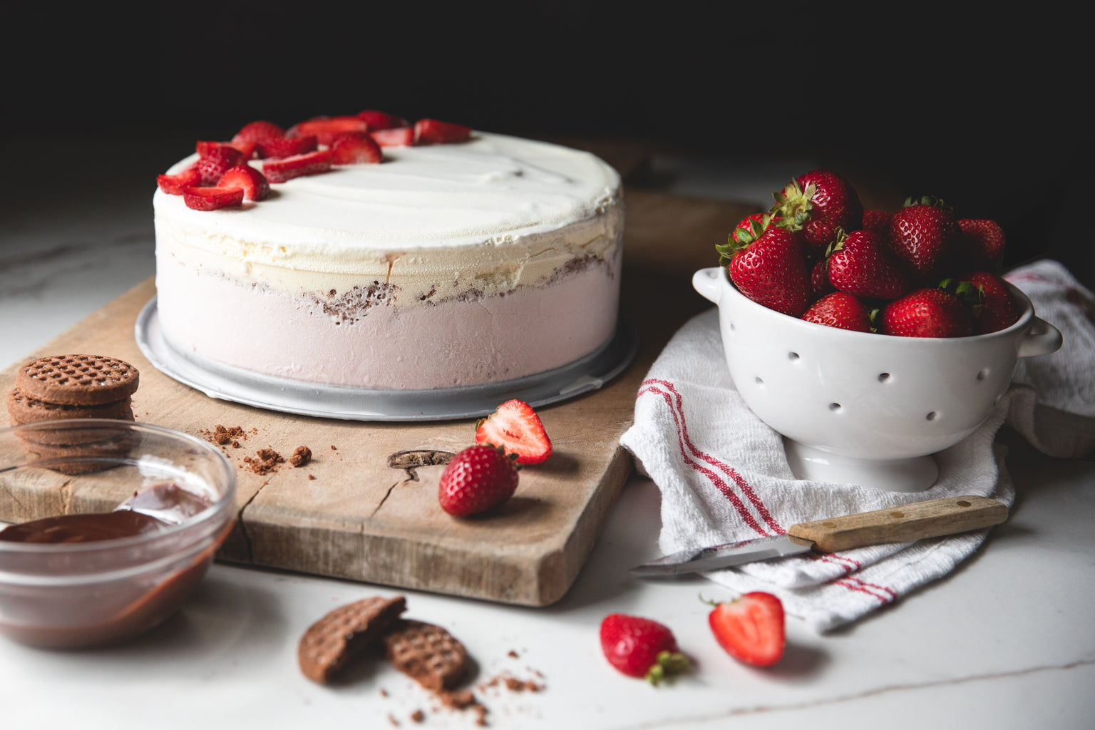 Strawberry ice cream cake topped with sliced strawberries on a wooden board, beside a colander of fresh strawberries, Einkorn Crispy Cocoa Cookies, and chocolate sauce
