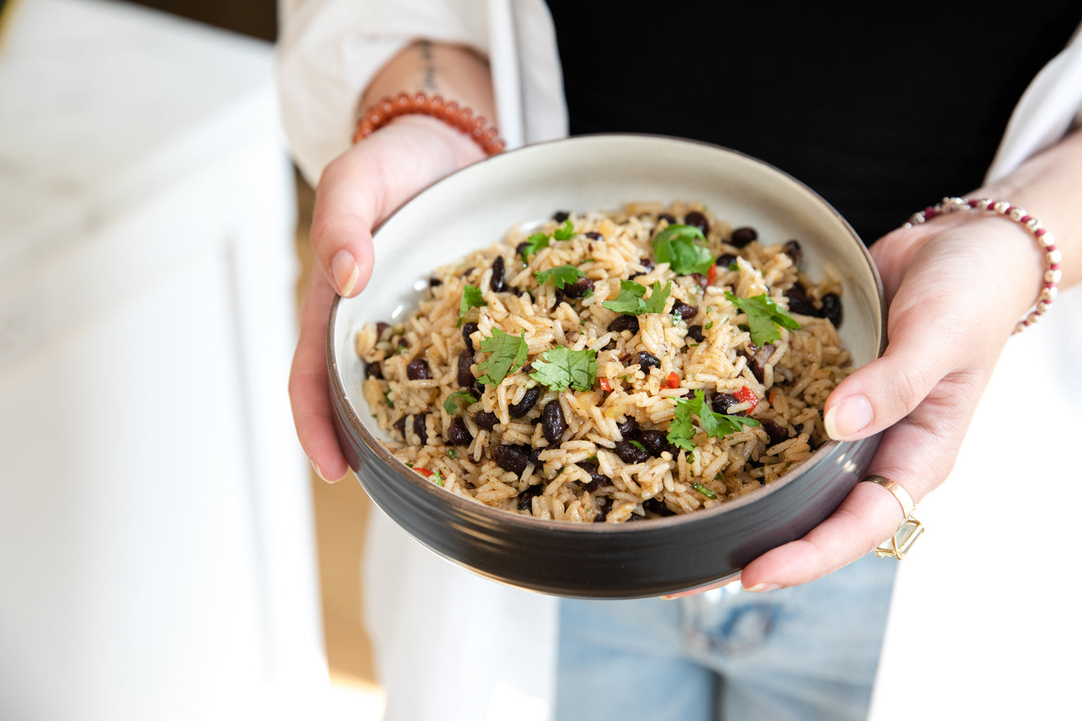 Person holding a bowl of black beans and rice garnished with fresh herbs in a bright kitchen