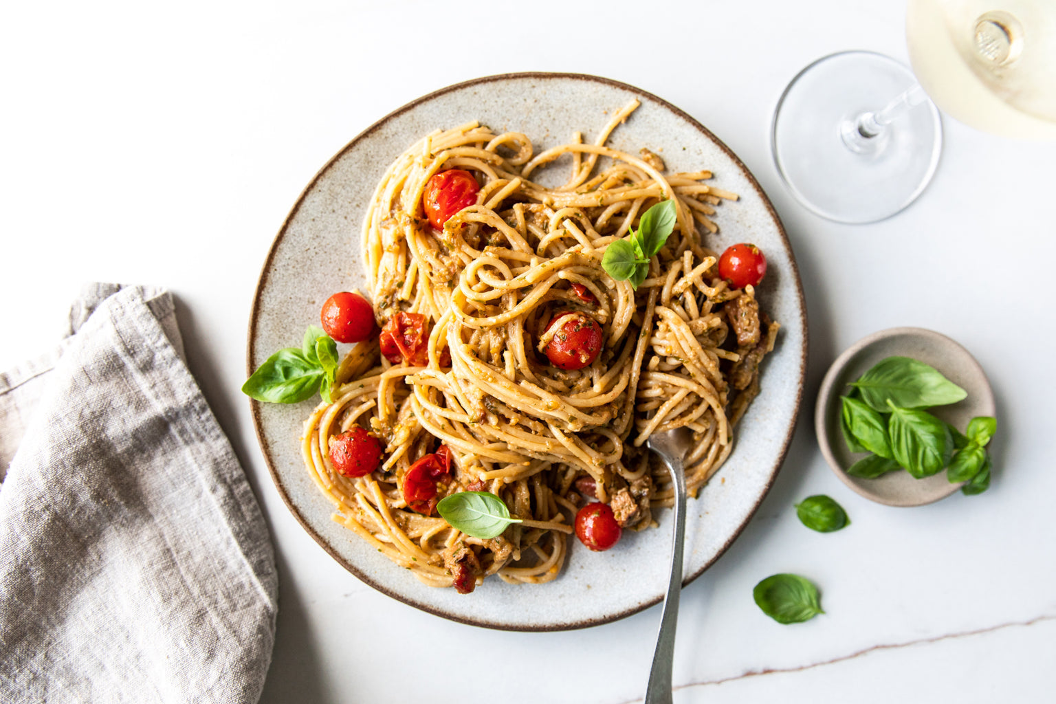 Plate of spaghetti with eggplant sauce and cherry tomatoes, garnished with basil on a white table beside a linen napkin and a glass of white wine