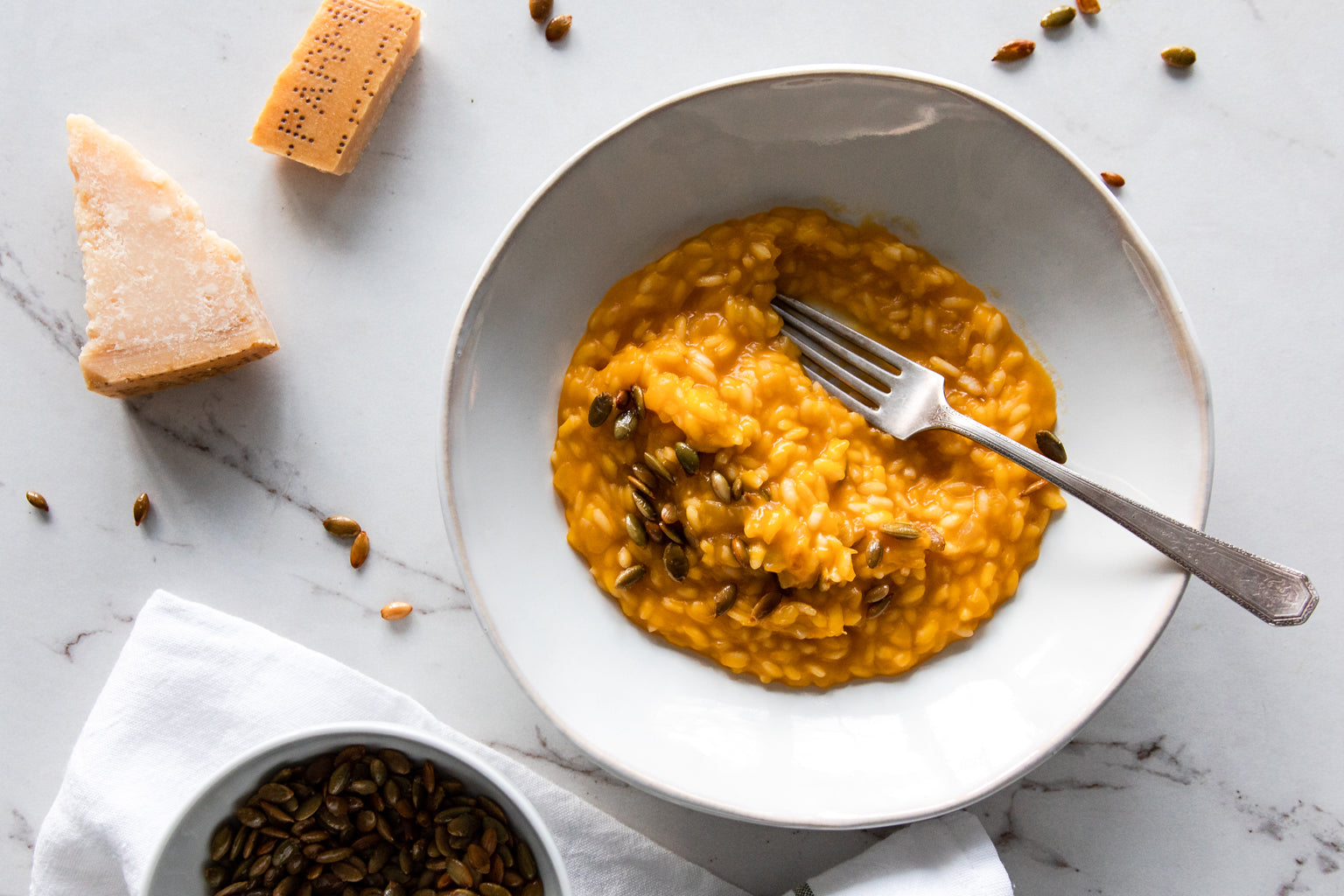Butternut squash risotto served in a white bowl with toasted pumpkin seeds, fork, and Parmesan wedge on a marble surface