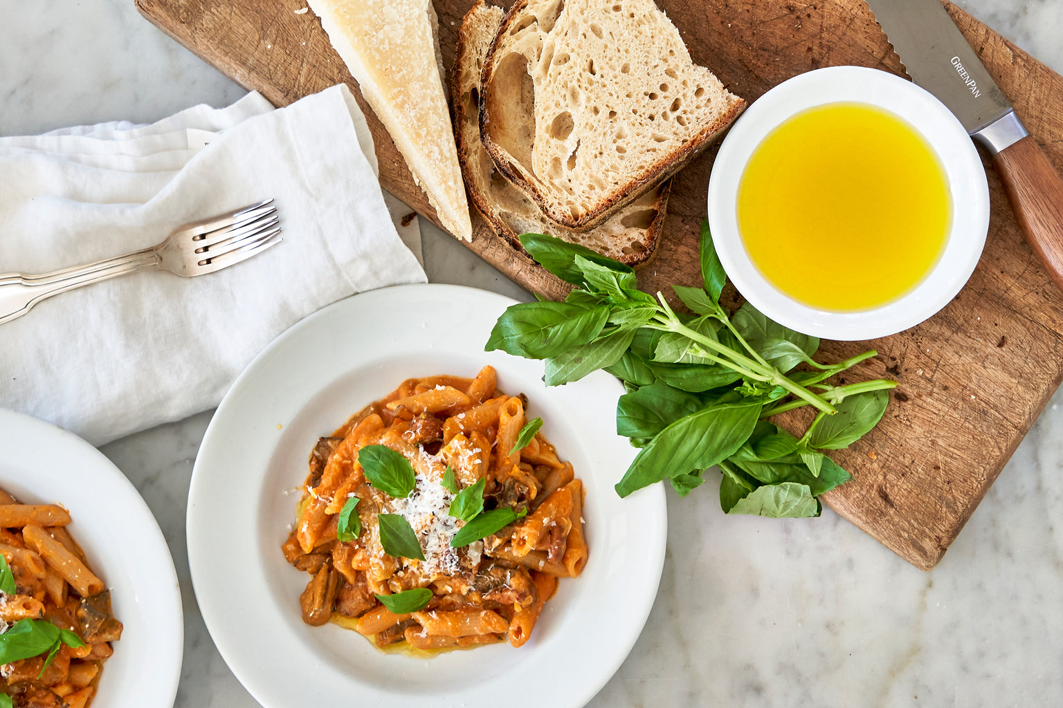 Penne in creamy butternut squash vodka sauce served on white plates with fresh basil, crusty bread, a wedge of cheese, and a bowl of olive oil on a marble surface