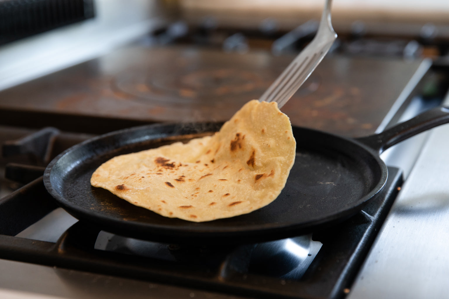 Einkorn chapatti being lifted with a fork from a hot cast-iron skillet on a gas stove