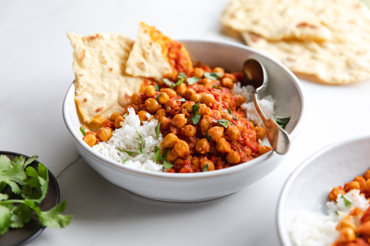 Bowl of vegan chickpea curry served over white rice with cilantro and torn flatbread on a light tabletop