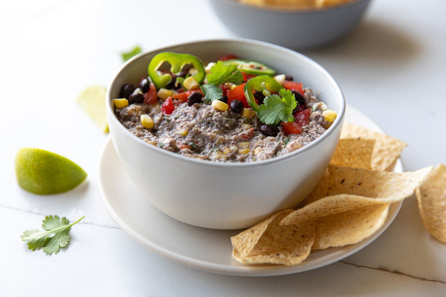 Bowl of black bean dip topped with corn, diced tomatoes, sliced jalapeño and cilantro, served with tortilla chips on a white plate