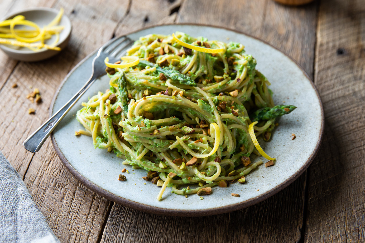 Plate of einkorn spaghetti tossed in creamy asparagus and ricotta sauce with lemon zest and chopped pistachios on a rustic wood table