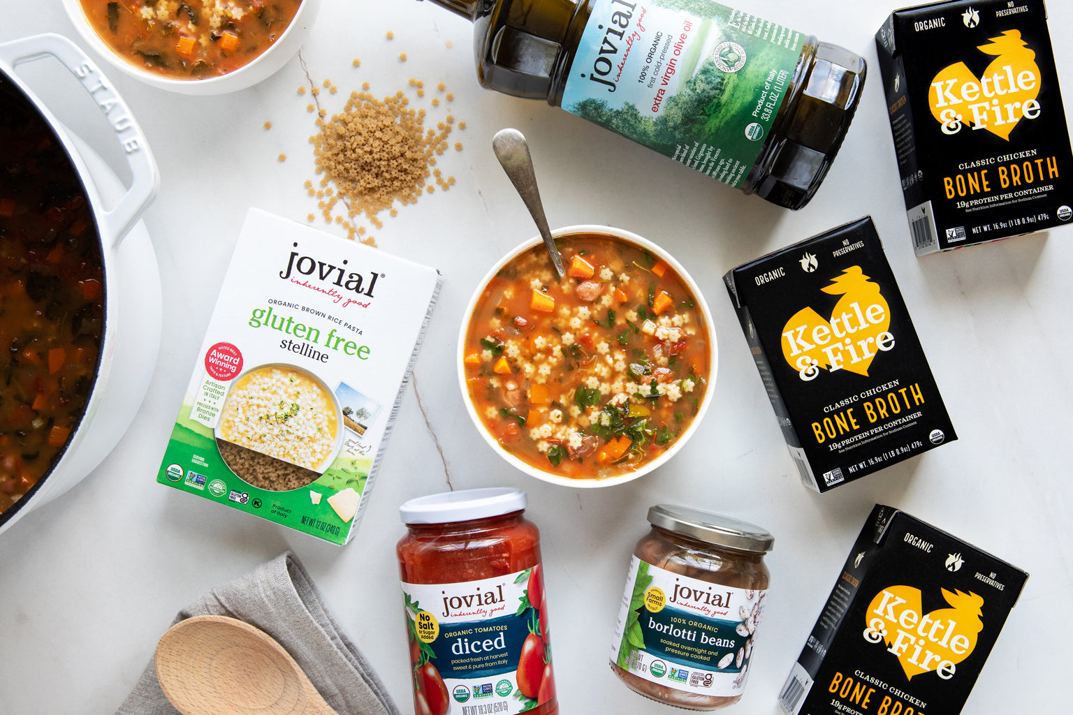 Bowl of stelline minestrone soup on a marble countertop surrounded by Jovial jars and pasta box, Kettle & Fire bone broth cartons, and a serving pot