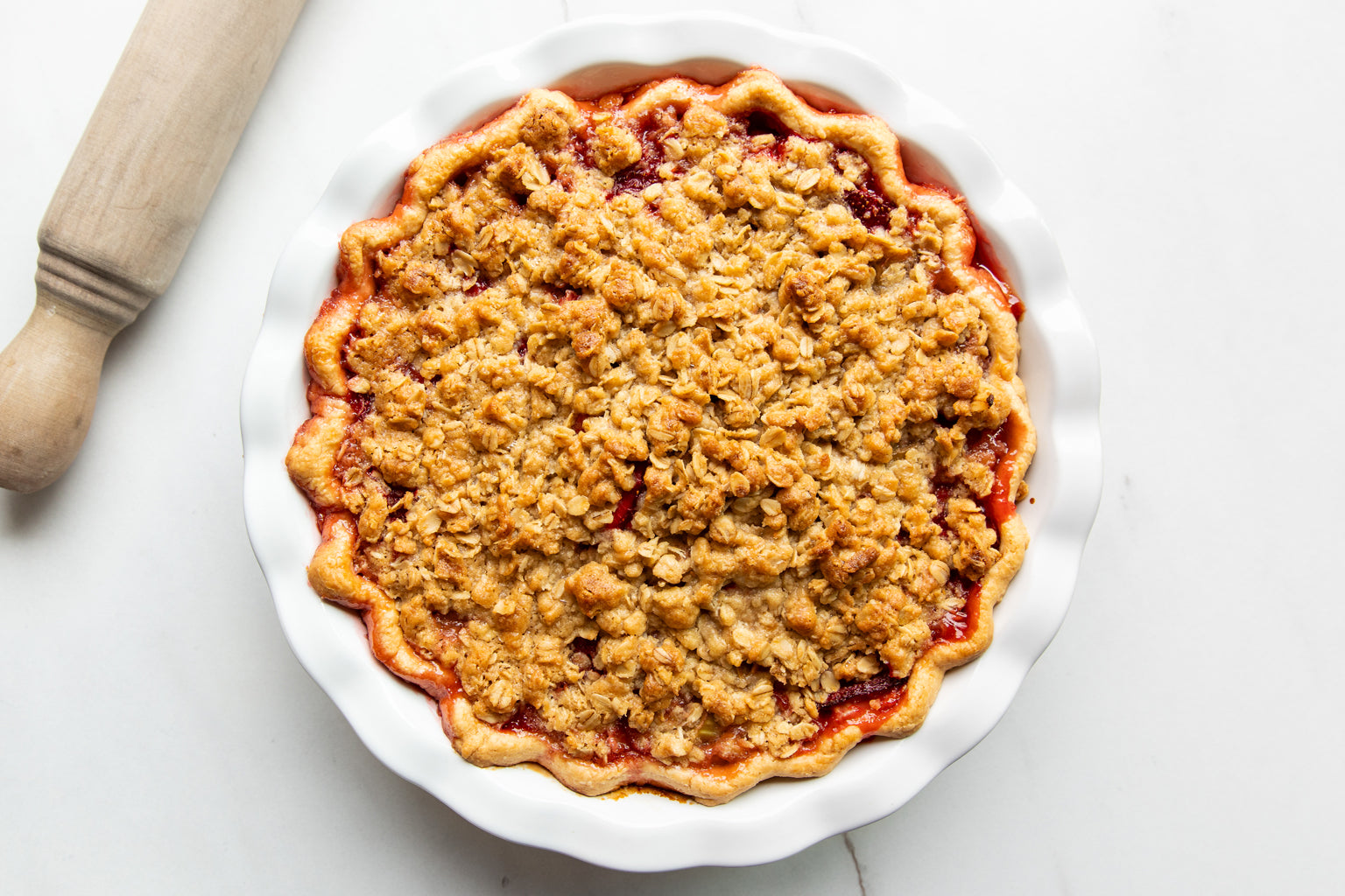 Top view of strawberry rhubarb pie with Einkorn oat crumble topping in a white fluted dish on a light marble surface with wooden rolling pin