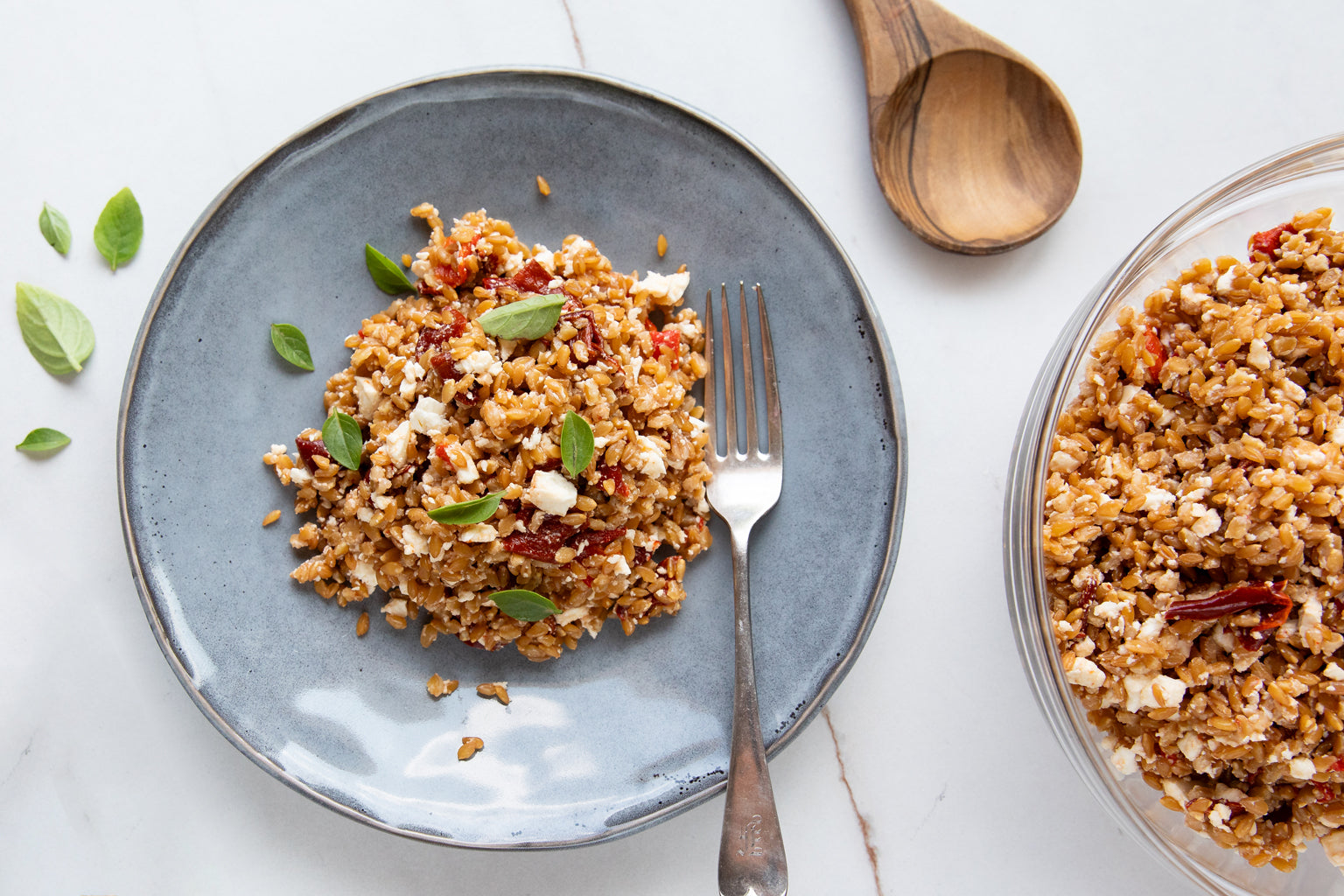 Roasted red pepper and Einkorn wheat berry salad on a blue plate with basil leaves and a fork, bowl of salad and wooden spoon on a marble surface