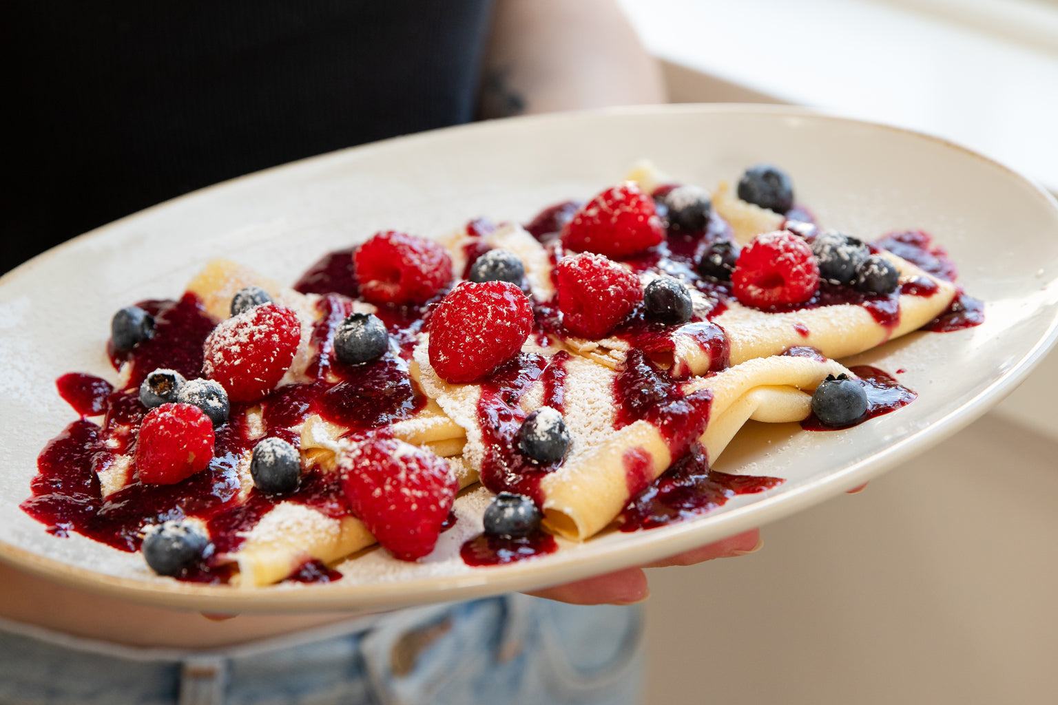 Einkorn crêpes folded on a plate with summer berry sauce, raspberries, blueberries and powdered sugar, person holding plate