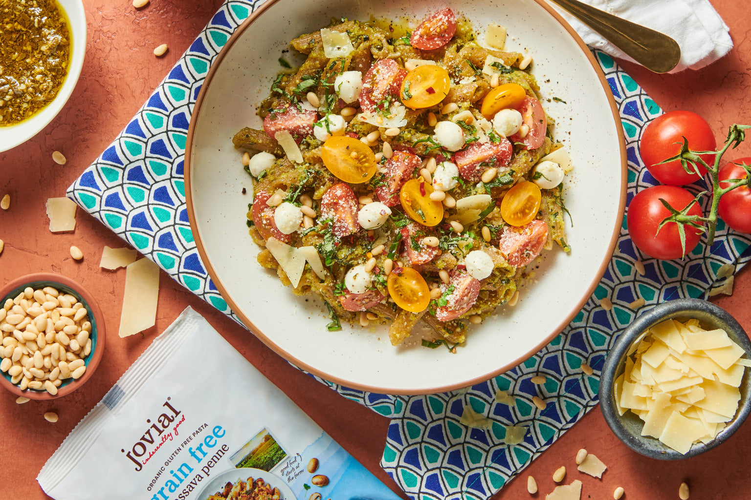 Bowl of Caprese pesto pasta with cherry tomatoes, mozzarella, pine nuts, and shaved Parmesan on a patterned napkin beside a Jovial grain-free pasta package