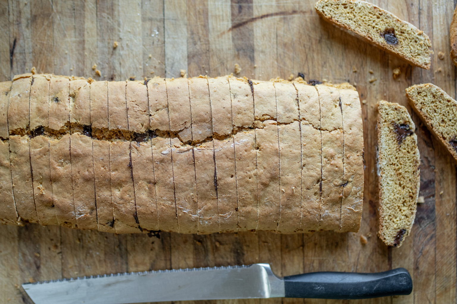 Top-down view of Einkorn biscotti loaf sliced into thin pieces with chocolate bits and cracked crust on wooden board