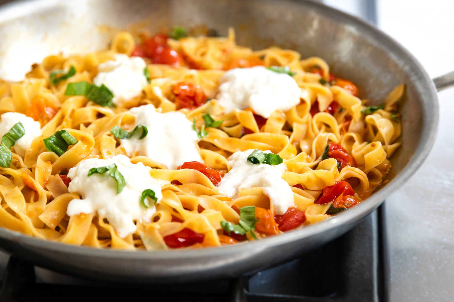Tagliatelle tossed with blistered cherry tomatoes, dollops of burrata and torn basil in a steel skillet on stovetop