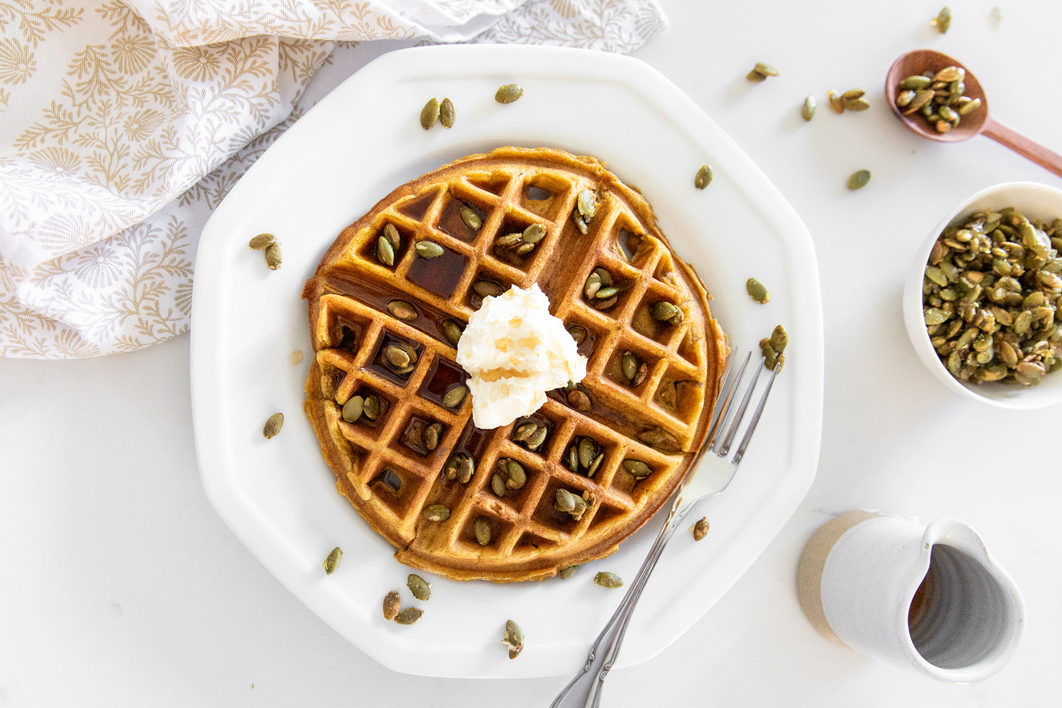 Pumpkin Einkorn waffle topped with whipped cream and pepitas on a white plate with a fork and syrup pitcher nearby