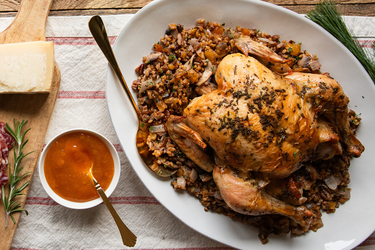 Roasted whole chicken on a platter of wheat berry stuffing with a bowl of sauce and rosemary on a striped linen
