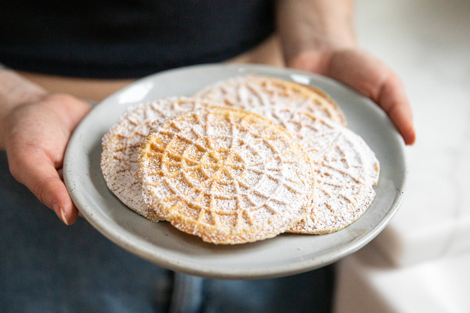 Three thin Einkorn pizzelle cookies dusted with powdered sugar on a ceramic plate held by hands