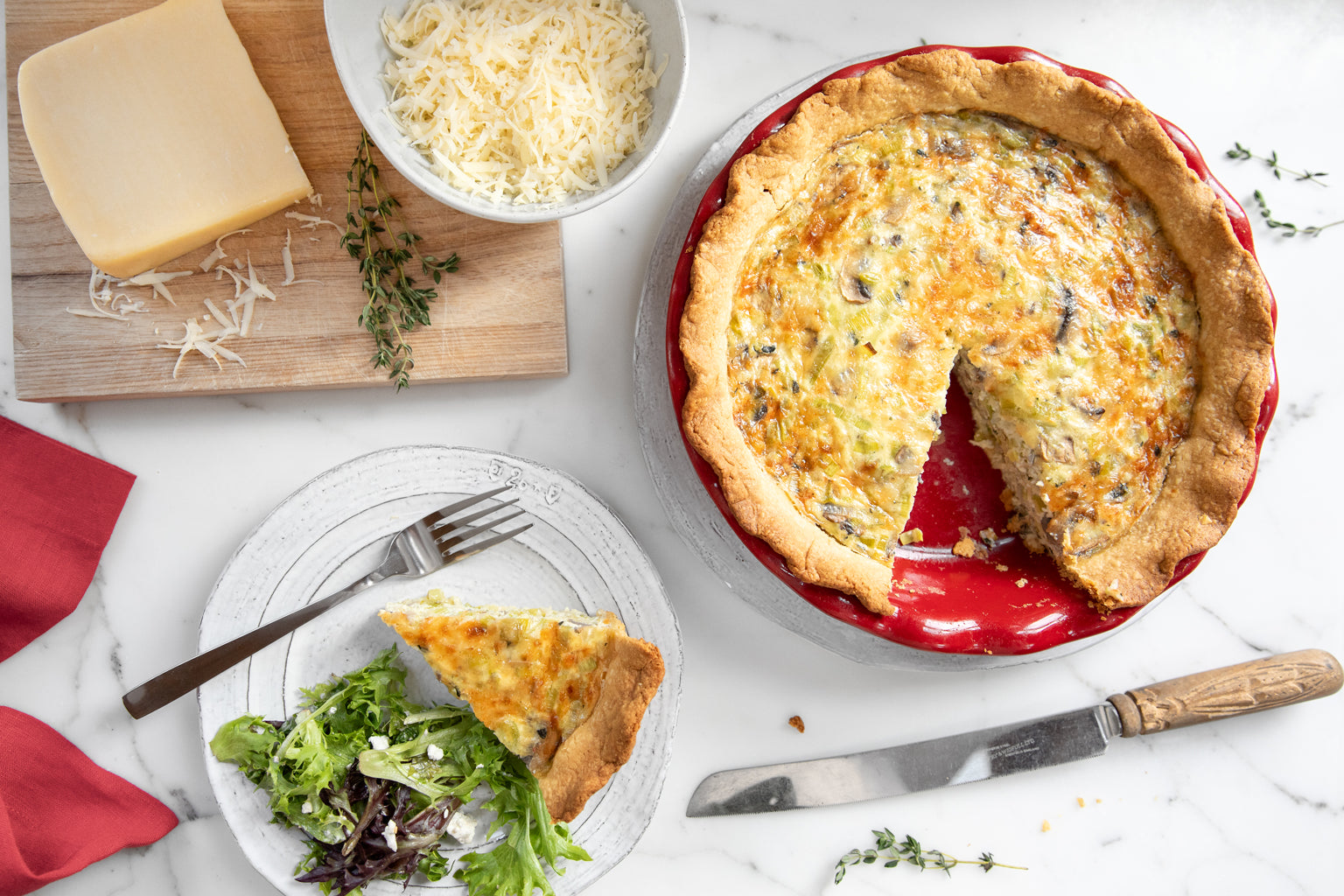 Leek and mushroom Einkorn quiche in a red pie dish with one slice removed, plated slice with mixed greens on a marble table