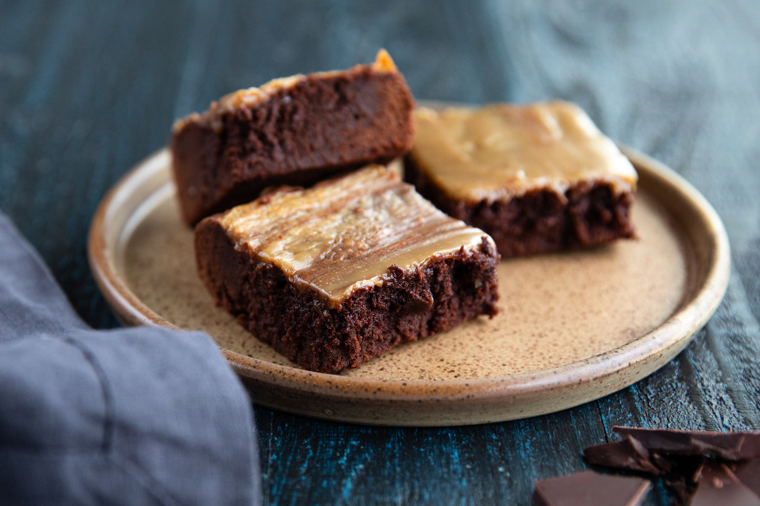 Close-up of three fudgy einkorn halva brownies with glossy halva topping on a speckled ceramic plate on a dark wood table