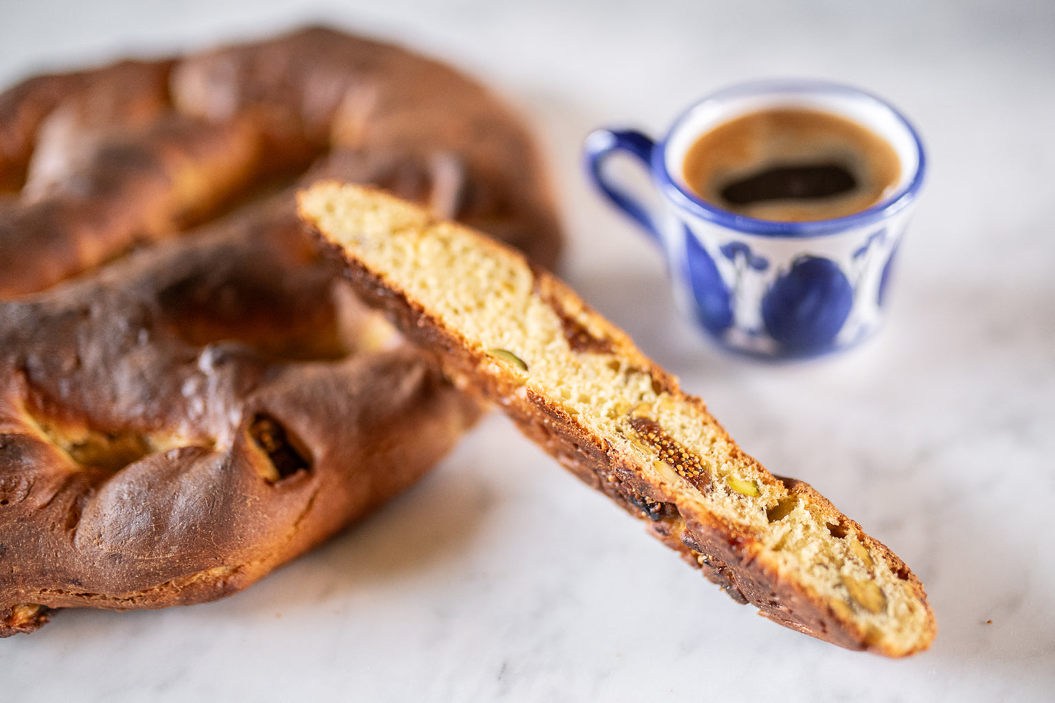 Slice of Einkorn breakfast bread showing figs and pistachios, with whole loaf and blue espresso cup on marble