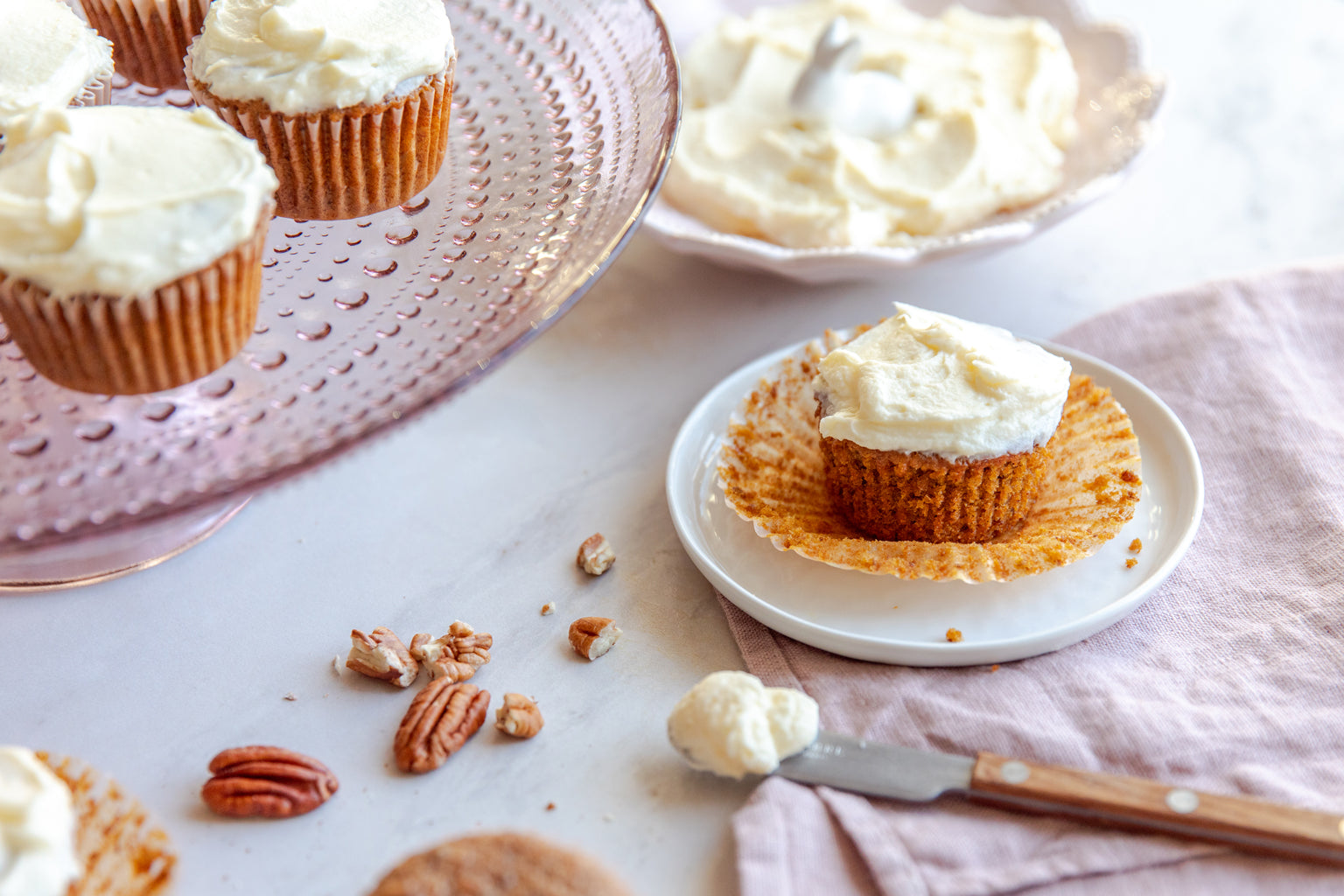 Einkorn carrot cupcake with cream cheese frosting sits on a small white plate beside a pink glass stand and pecans