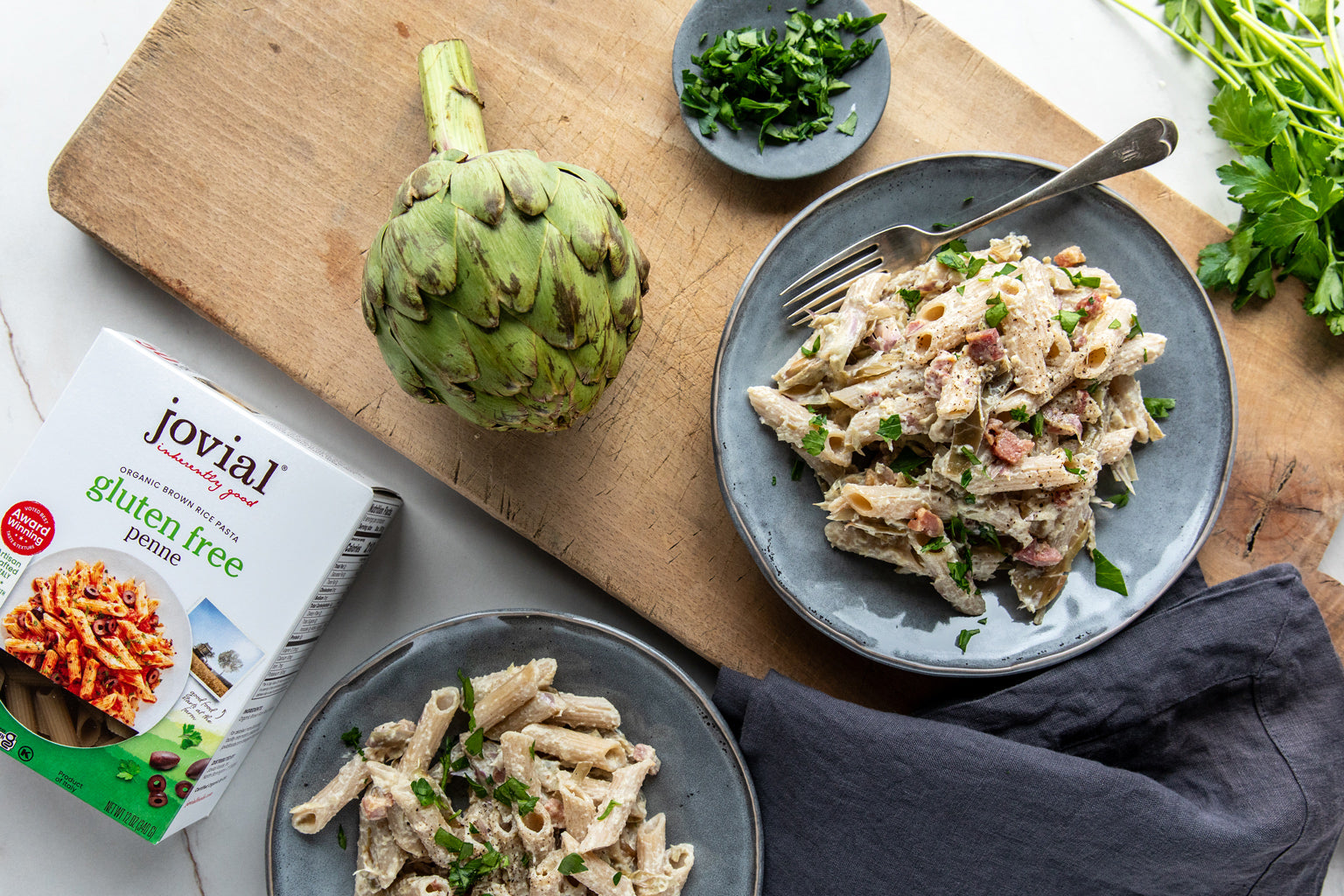 Two bowls of artichoke penne with crispy pancetta on a wooden board beside a gluten free brown rice penne box and artichoke