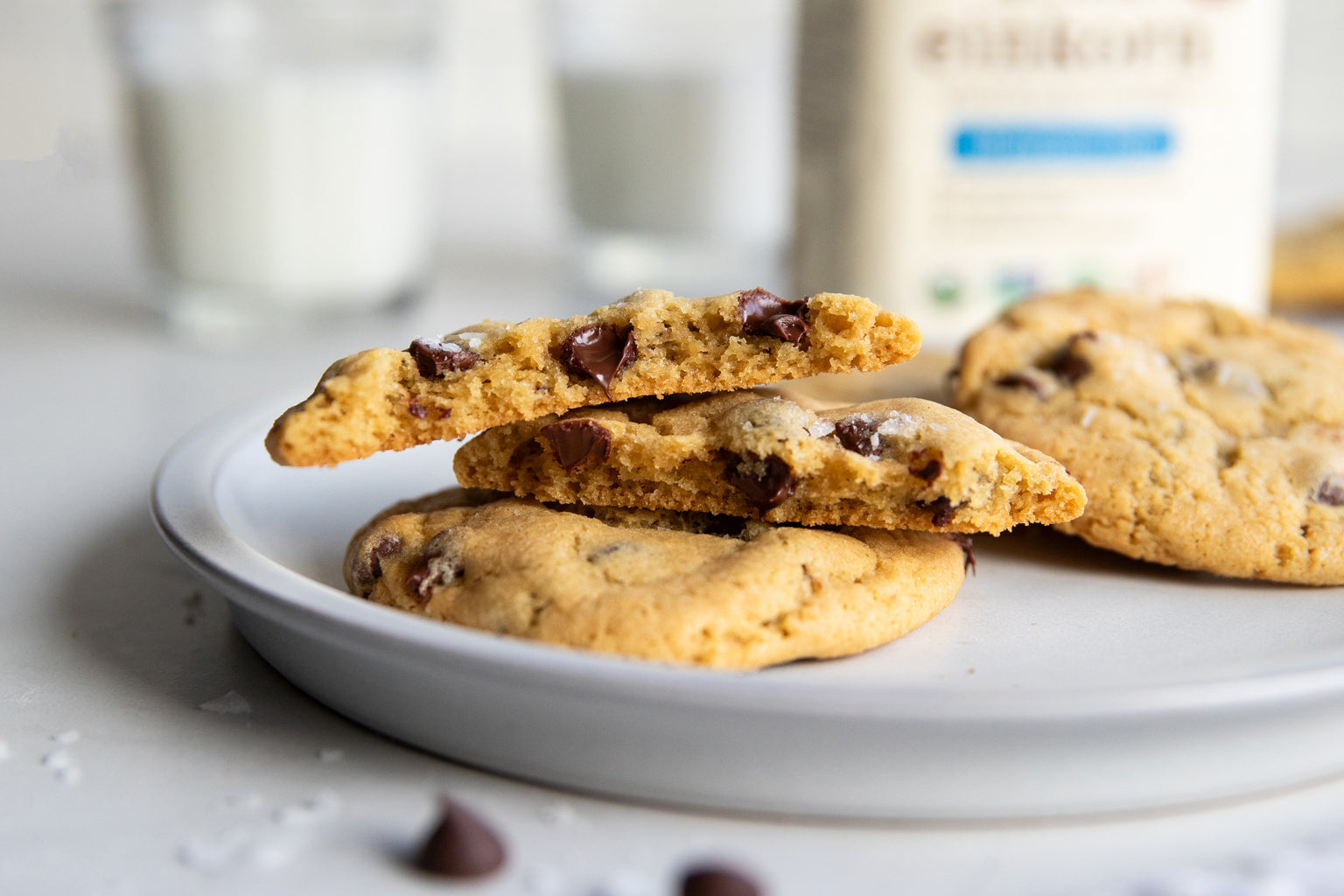 Close-up of Einkorn chocolate chip cookies on a plate with a broken cookie showing chocolate chips, blurred milk in background