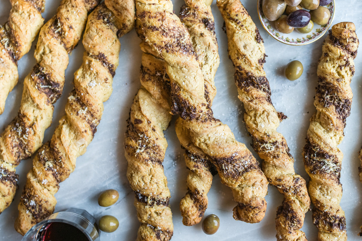 Overhead view of Einkorn grissini twisted with olive tapenade swirls on a marble surface with olives and a small bowl