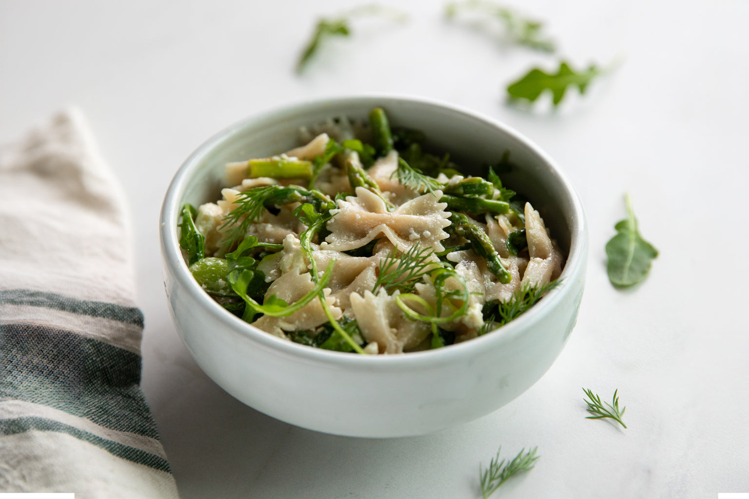 Bowl of brown rice farfalle pasta salad with asparagus, arugula, and dill on a white surface