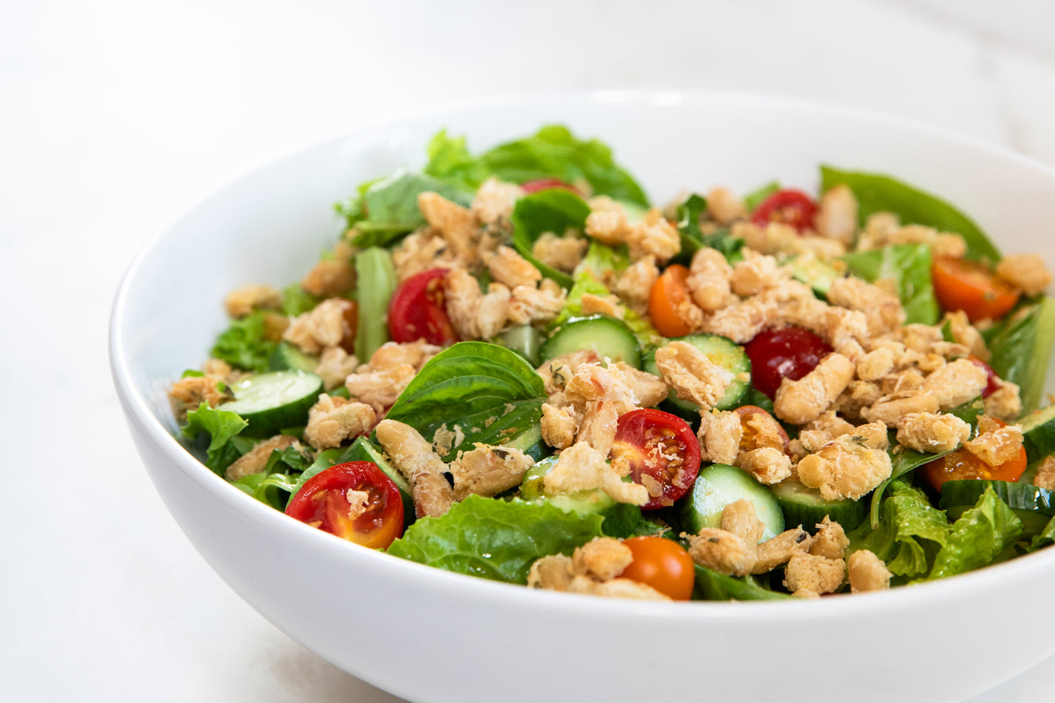 Bowl of salad topped with crispy cannellini beans, cherry tomatoes, cucumber, and basil on a white countertop