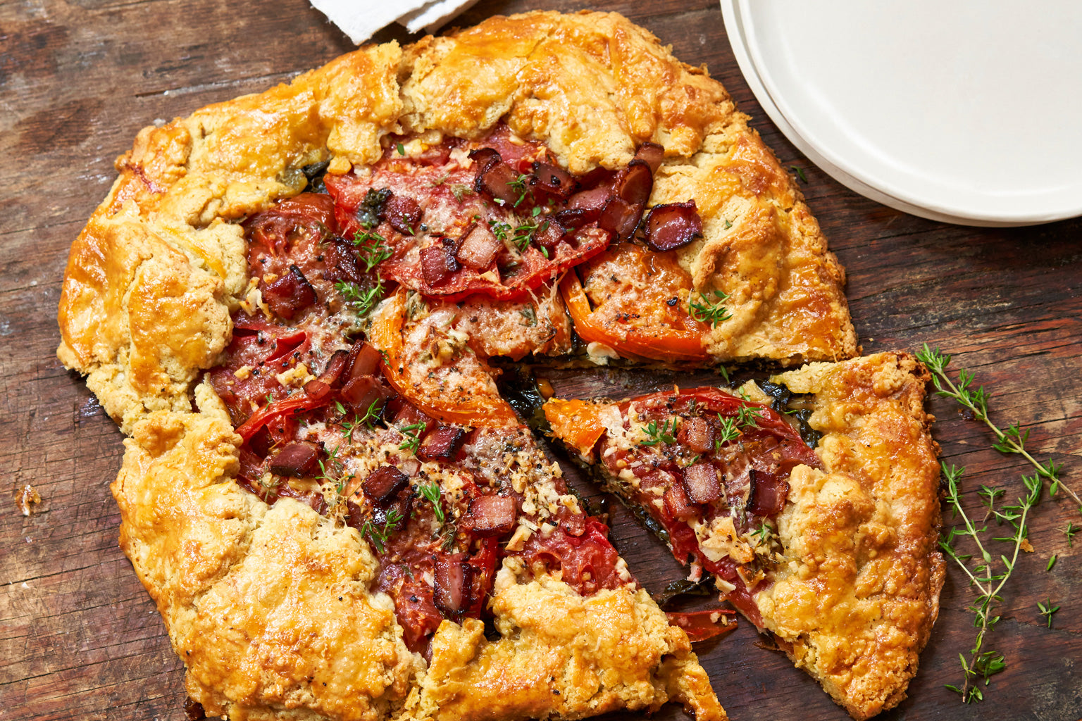 Einkorn BLT galette sliced on a wooden board showing tomatoes, bacon, melted cheese, and thyme sprigs