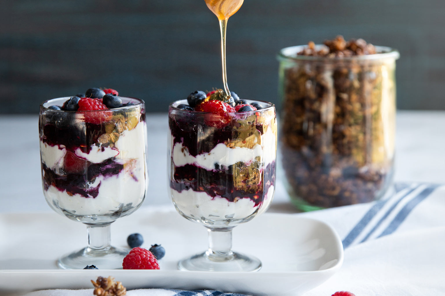 Two parfait glasses with berries, yogurt, and Einkorn wheat berry granola as honey is drizzled, jar of granola in background