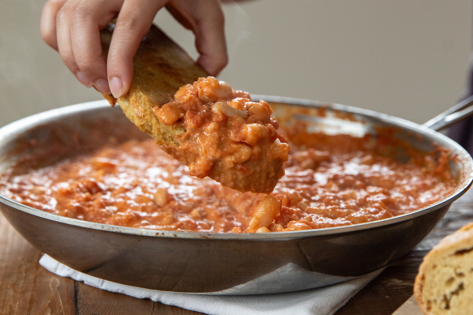 Hand dipping crusty bread into a skillet of creamy cannellini beans and tomatoes on a wooden table