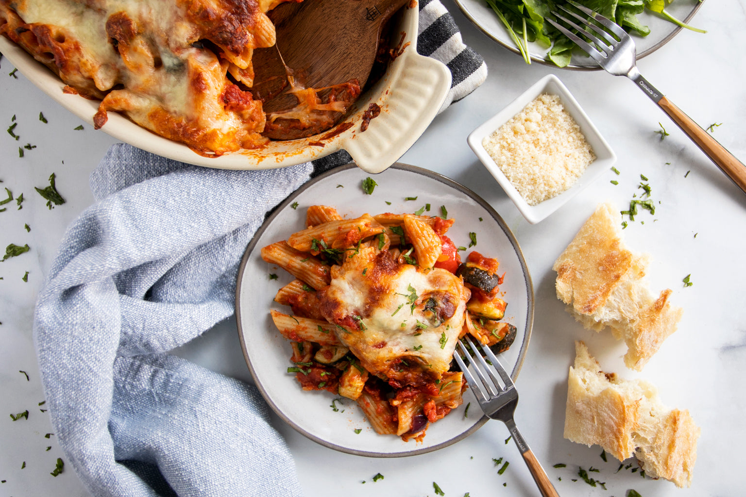 Baked penne with roasted vegetables and melted cheese on a plate next to a baking dish, grated cheese, torn bread, and a napkin