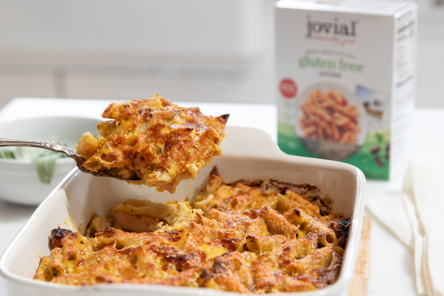 Spoon lifting butternut squash pasta al forno from white baking dish, golden cheese, gluten free pasta box blurred behind