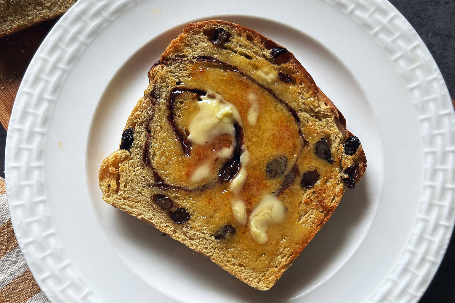 Top-down view of a slice of Sourdough Cinnamon Raisin Loaf showcasing a cinnamon swirl and melted butter on a white plate
