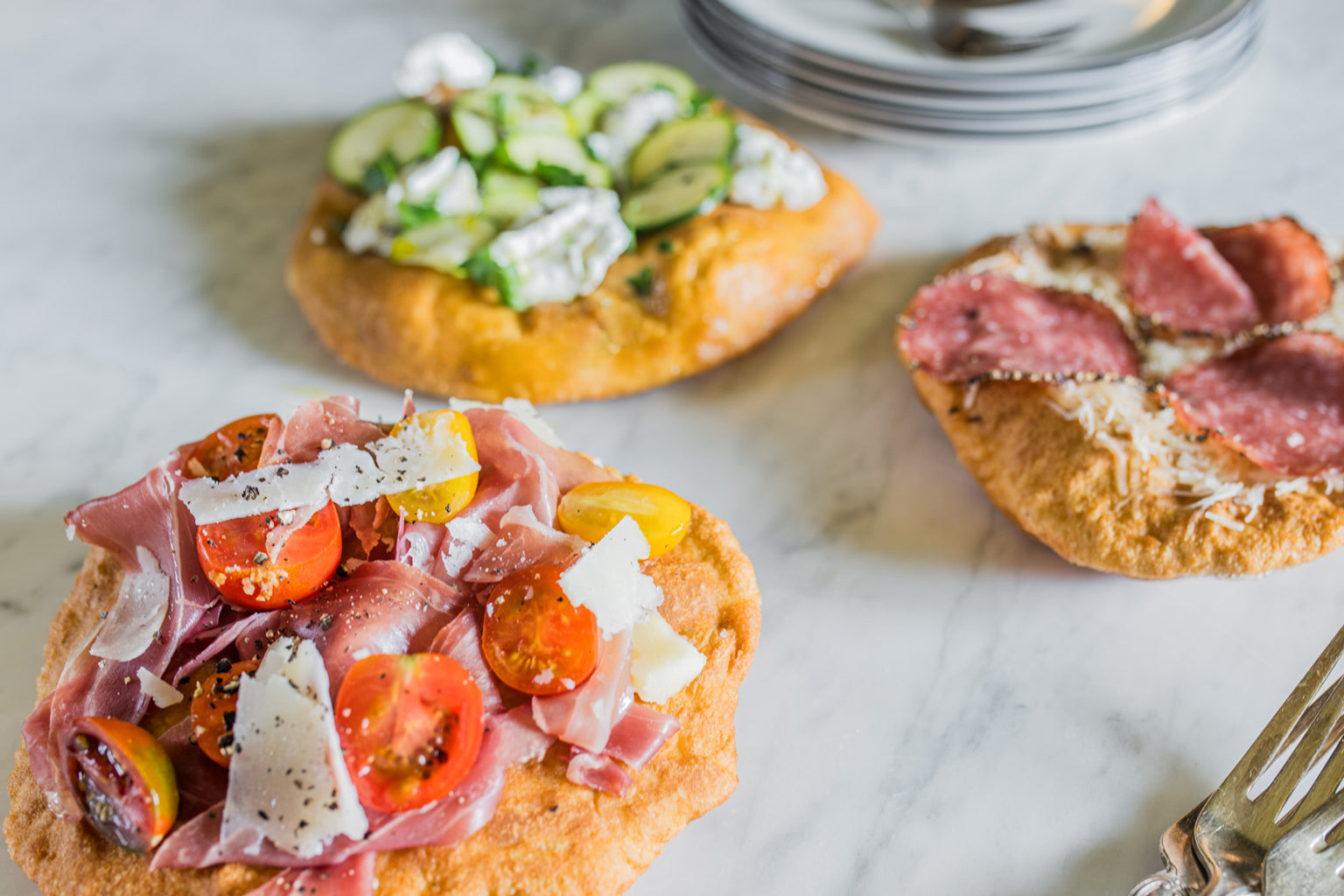 Three einkorn mini pizza fritta on a marble surface, topped with prosciutto and cherry tomatoes, zucchini with ricotta, and salami
