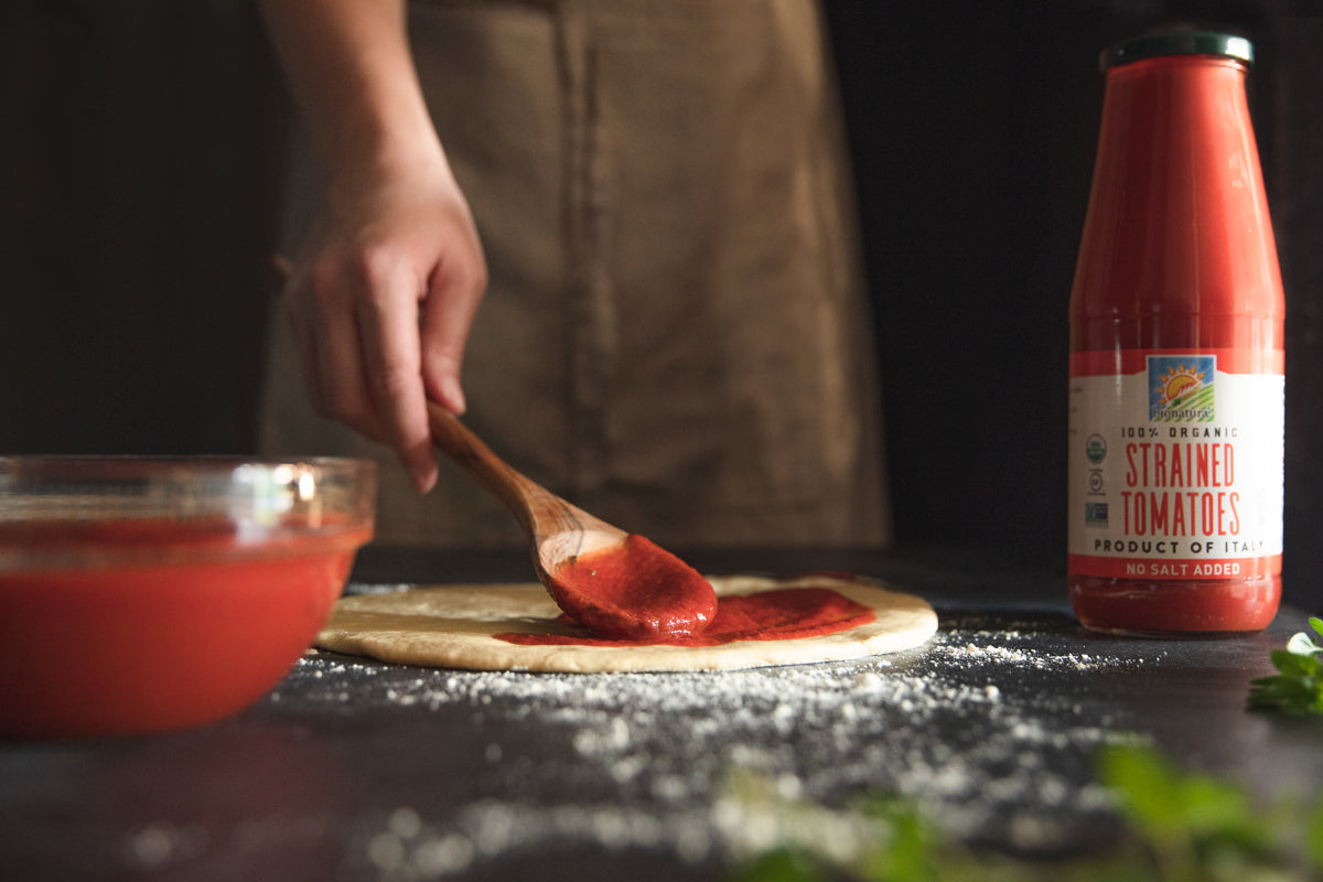 Person spreading tomato sauce on pizza dough, bowl and bottle of strained tomatoes on a flour-dusted counter