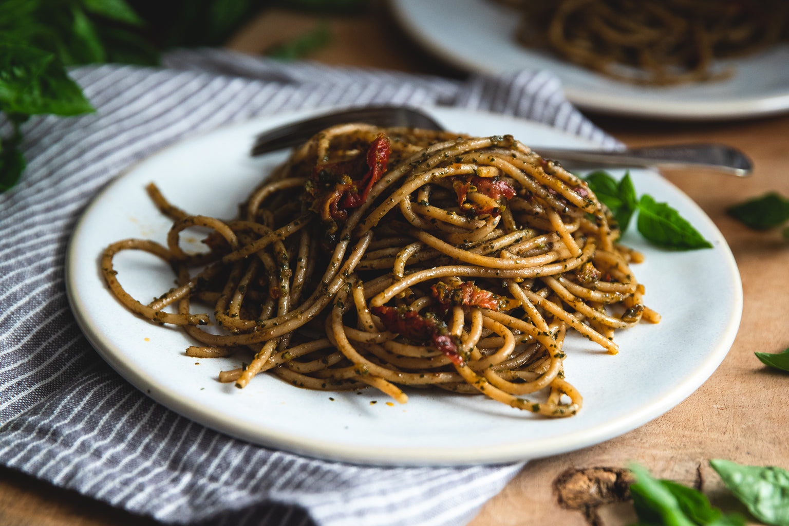 Plate of organic whole wheat spaghetti tossed with sundried tomato pesto, garnished with basil and a fork on a striped napkin
