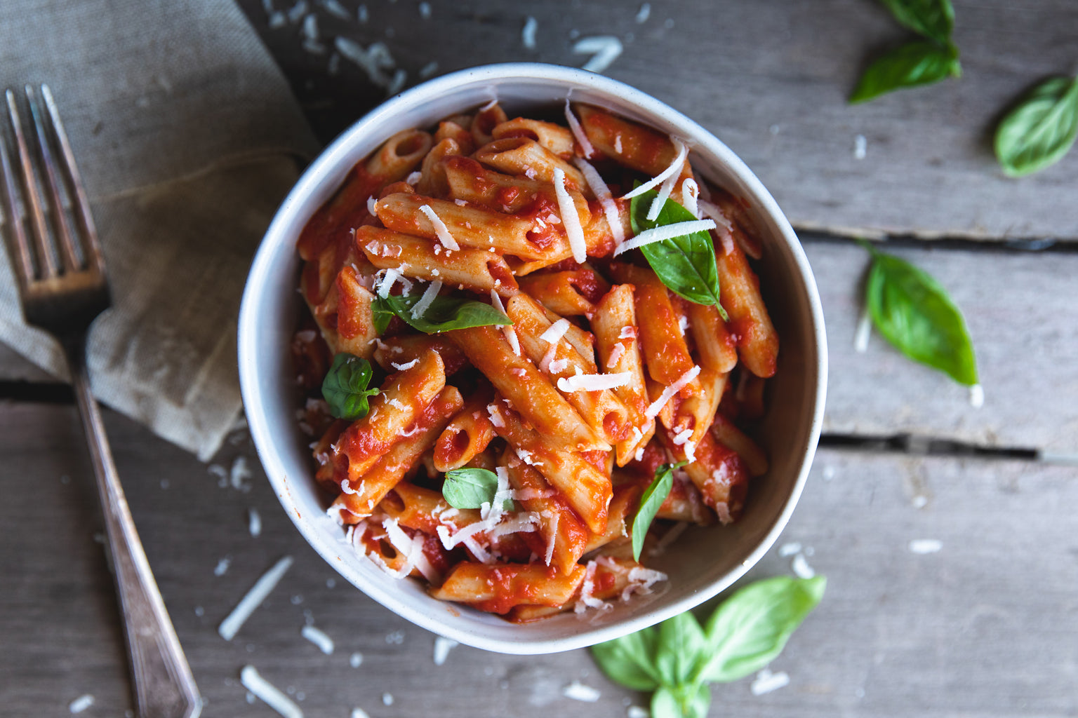 Bowl of penne rigate in tomato sauce with fresh basil and grated Parmigiano on a rustic wooden table