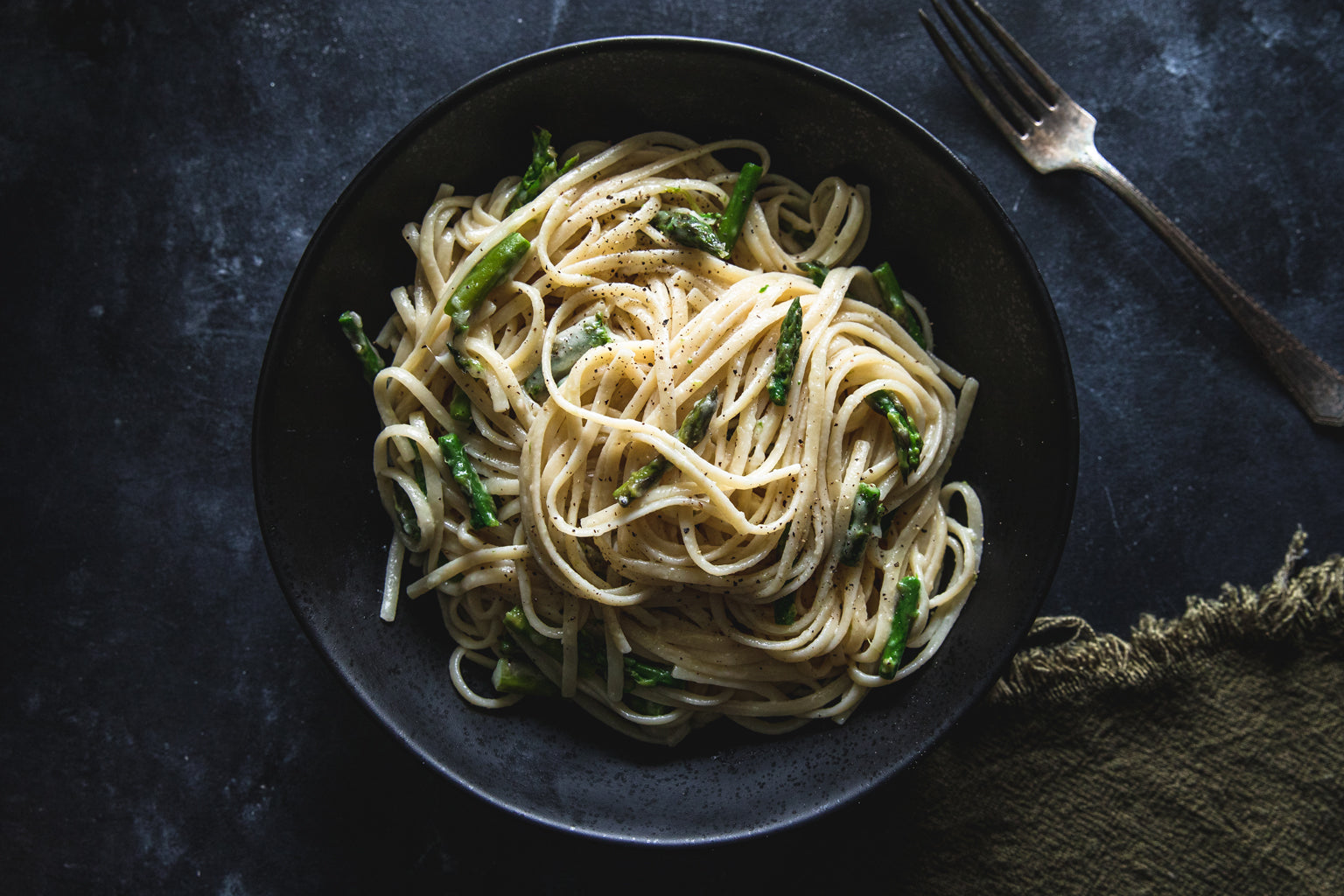 Bowl of linguine cacio e pepe tossed with asparagus and cracked black pepper on a dark tabletop with a fork at top right