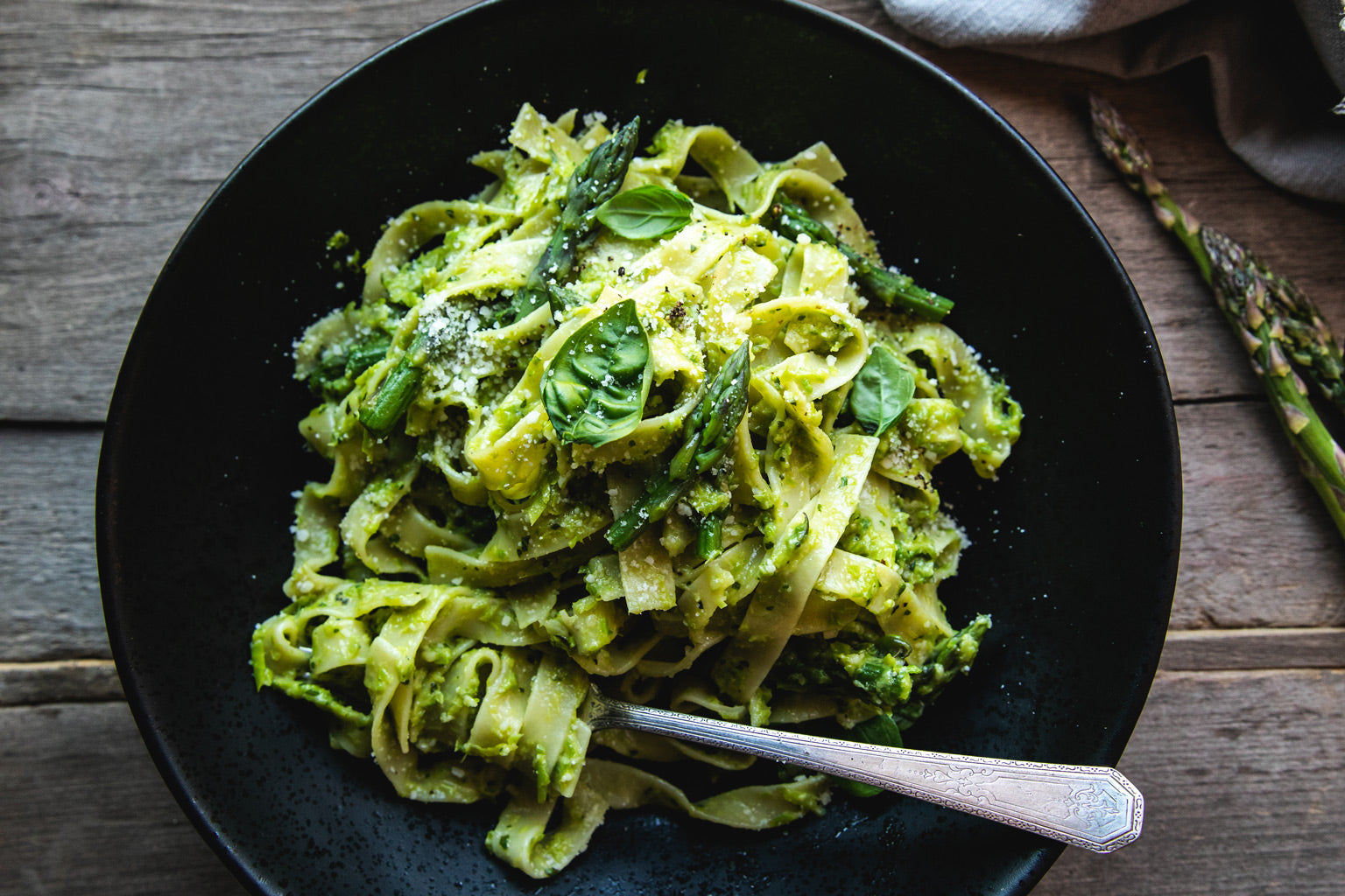 Egg tagliatelle tossed with asparagus pesto, asparagus tips, and grated Pecorino in a black bowl on a wooden table