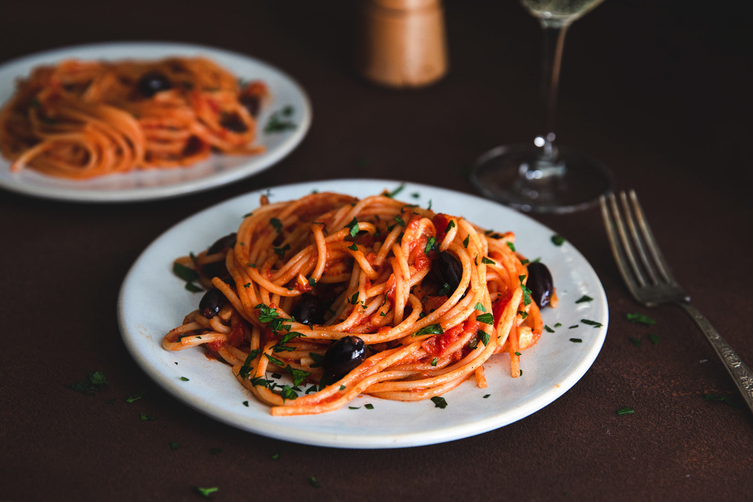 Spaghetti alla puttanesca served on a white plate with tomato sauce, black olives, and chopped parsley on a dark tabletop