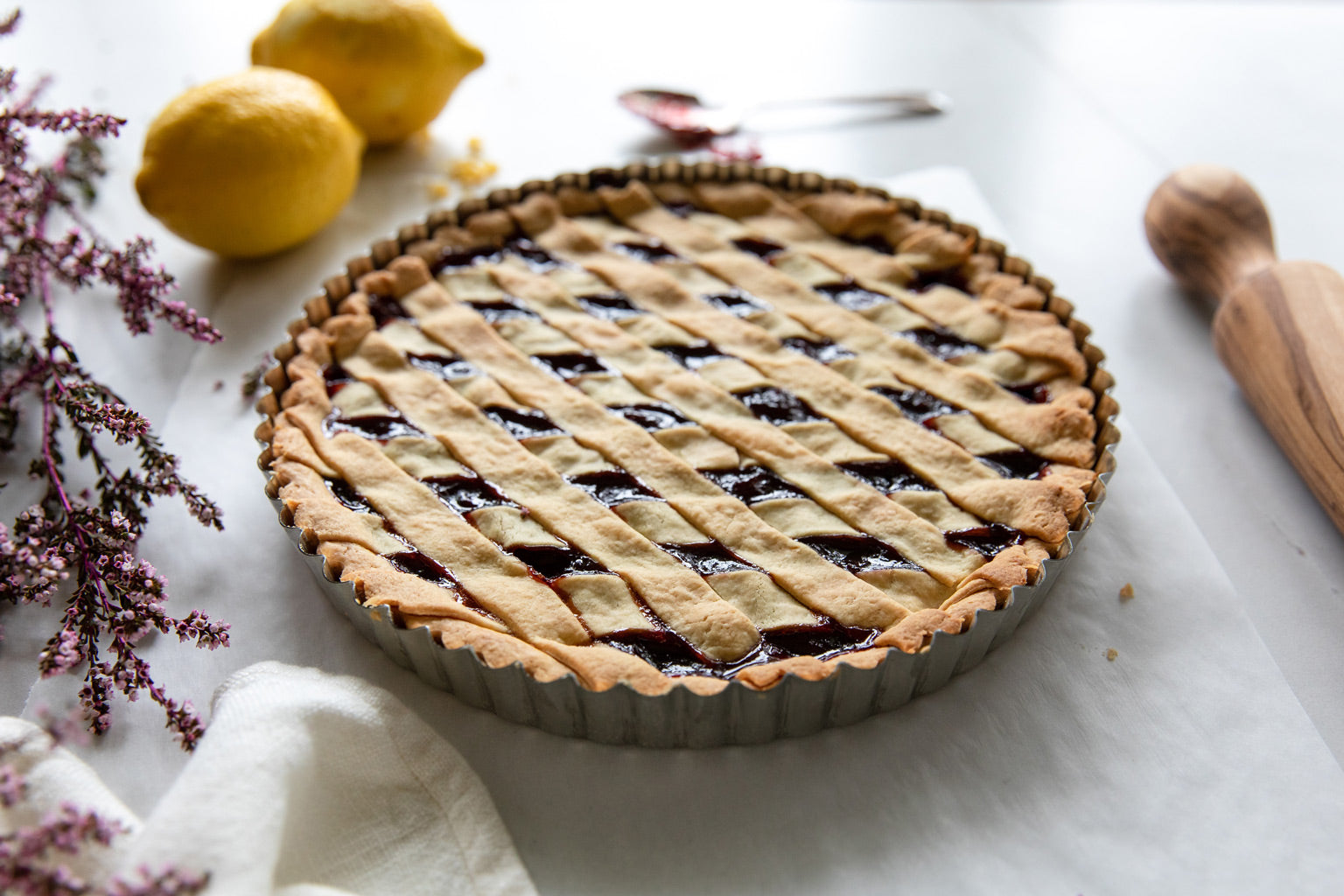 Sour cherry einkorn crostata cooling in a fluted tart pan on a white cloth with lemons, a rolling pin, and purple flowers