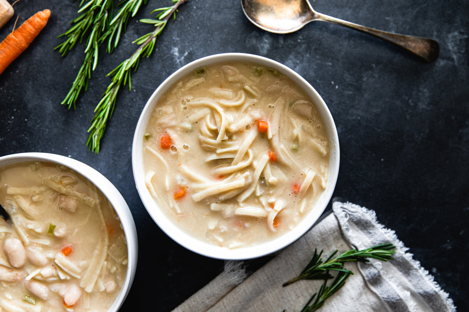 Two bowls of gluten-free cannellini bean soup with linguine on a dark tabletop, with rosemary sprigs, carrots, and a spoon